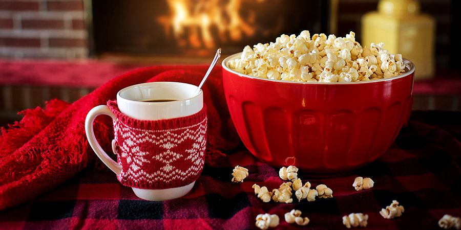 A bowl of popcorn and a mug of hot cocoa on a table near a lighted fireplace.